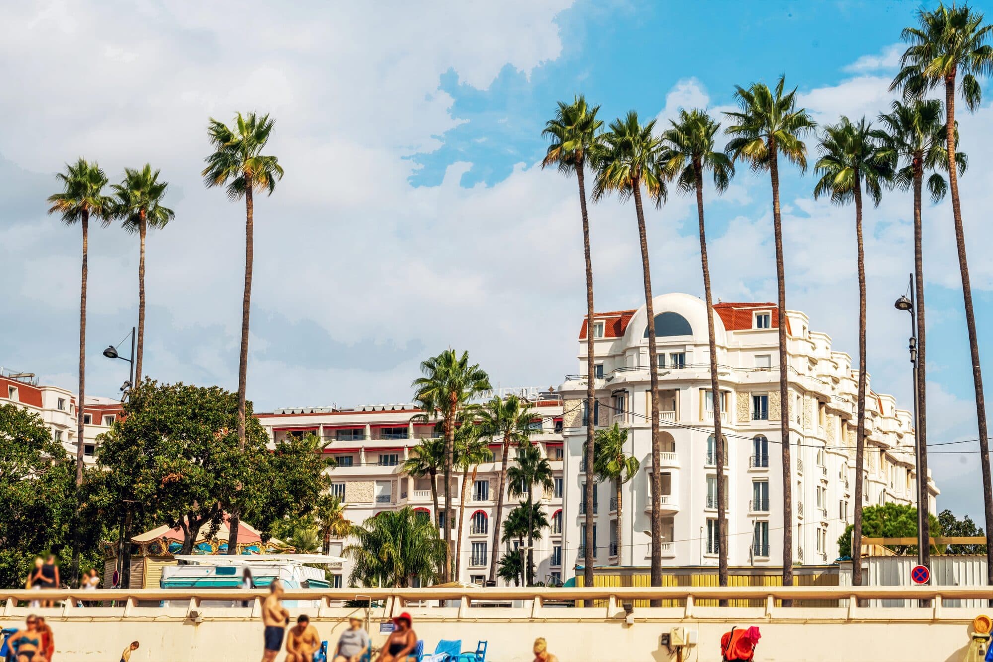 The white façade of a Croisette palace hotel in Cannes, framed by tall palm trees, seen from the beach