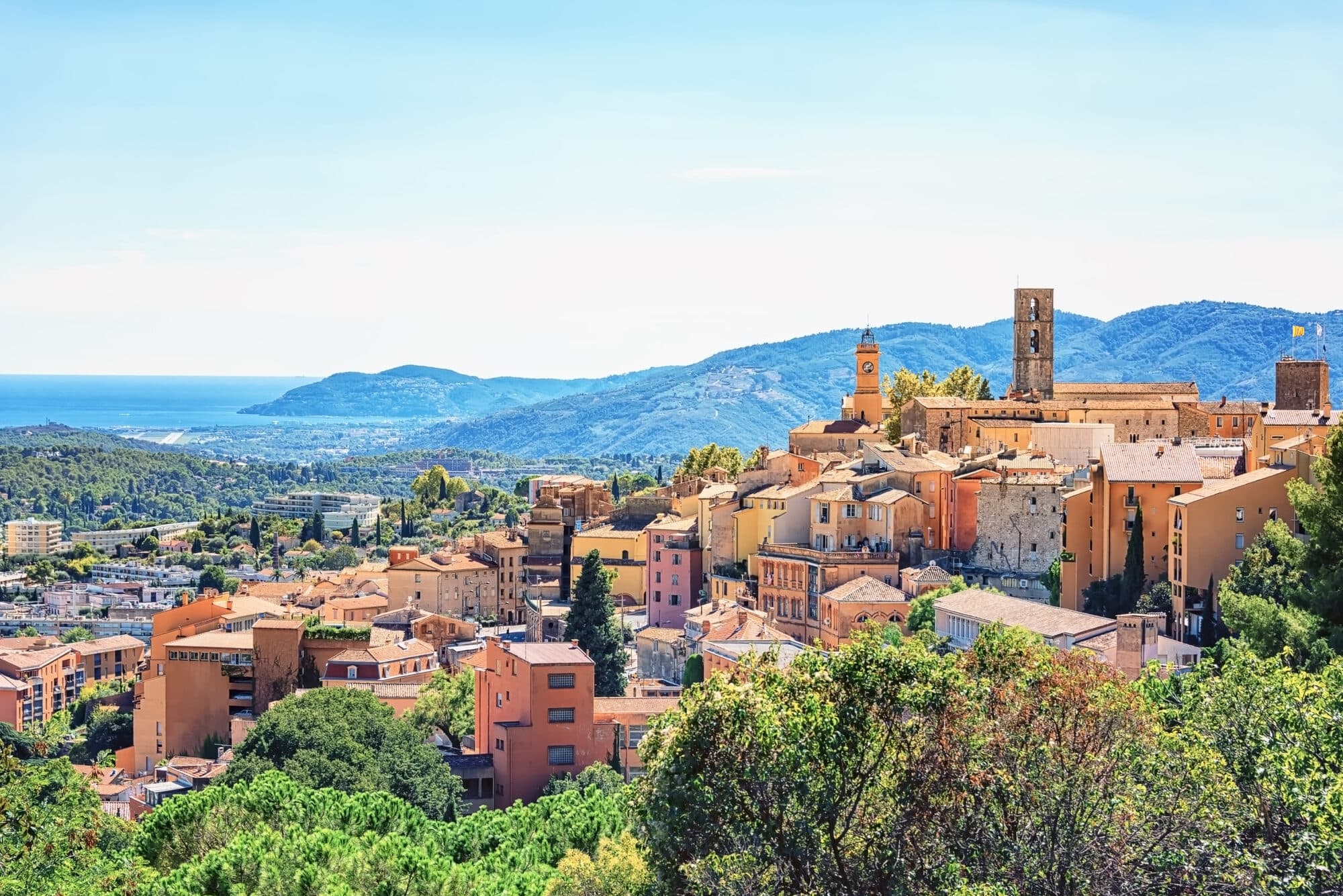 The old town of Grasse perched above the hills, its bell towers rising over the tiled roofs, the bay in the distance